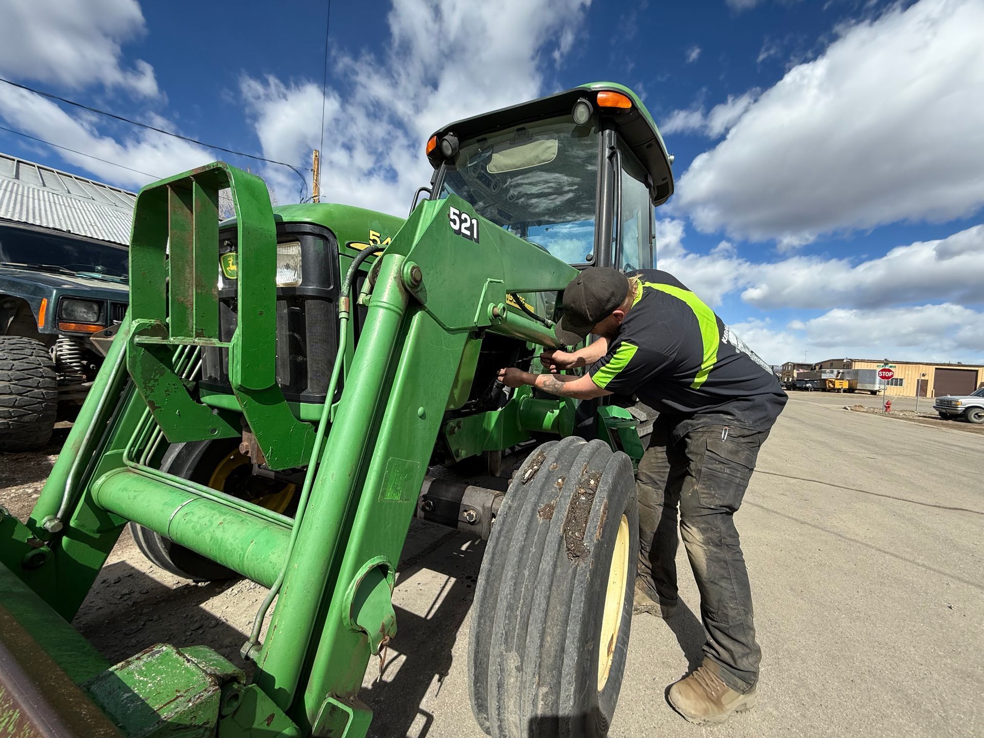 A mechanic in a high-visibility shirt inspects the engine of a large green tractor outdoors on a sunny day.