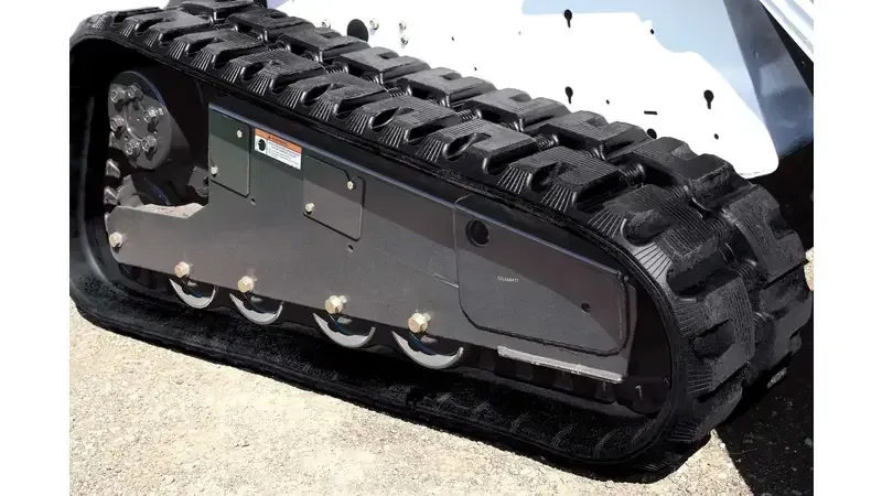 A close-up view of the black rubber track and steel undercarriage system of a compact track loader on a concrete surface.