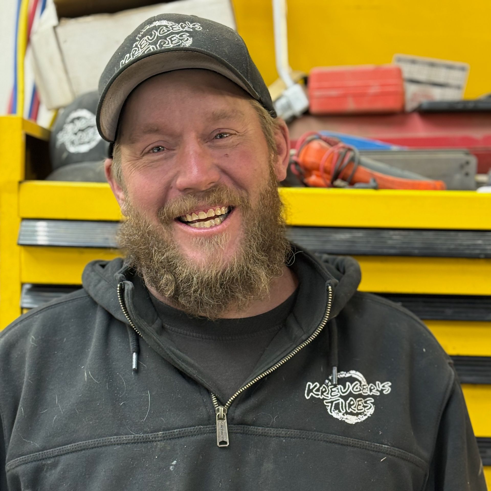 A smiling person with a beard and baseball cap stands in a workshop with yellow tool chests in the background.