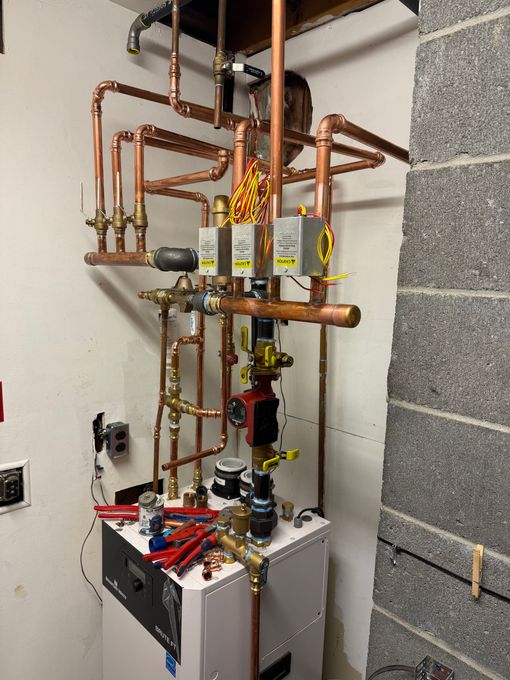 Copper plumbing and boiler equipment in a utility room, with tools on top and exposed pipes and valves.