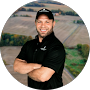 A man in a black cap and shirt with arms crossed, smiling against a backdrop of a rural field landscape.