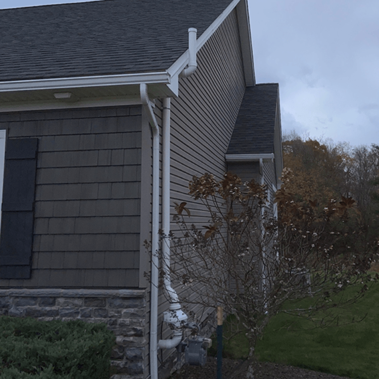 A white gutter downspout runs down the side of a house with tan siding and stone masonry, near a small, bare tree.
