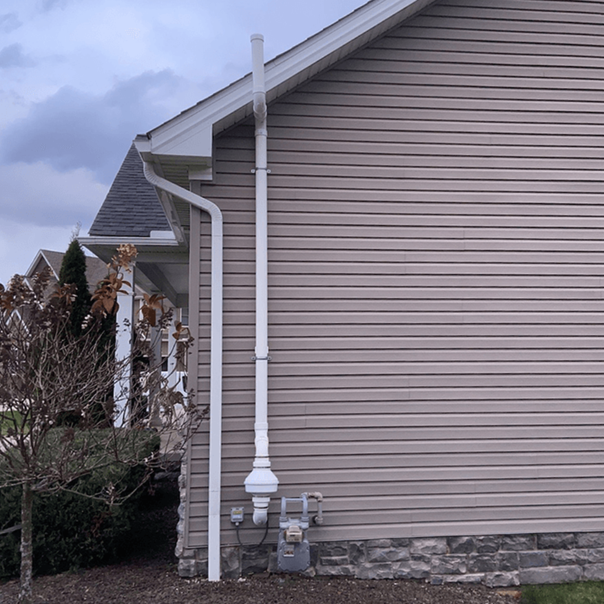 A white radon mitigation pipe runs vertically up the side of a tan vinyl-sided house next to a gas meter.