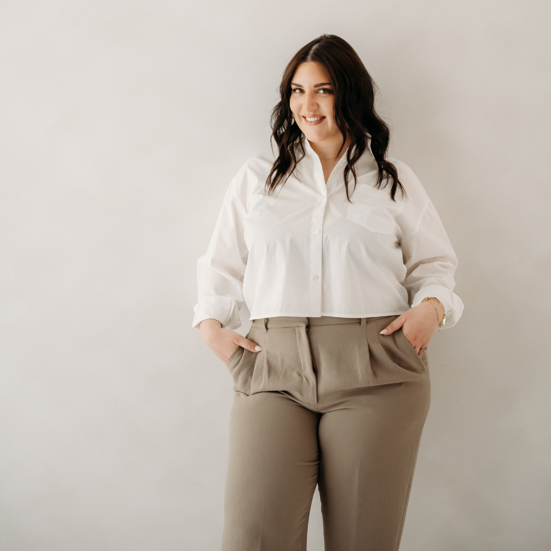Woman standing against a plain background, wearing a white shirt and beige pants, with hands in pockets