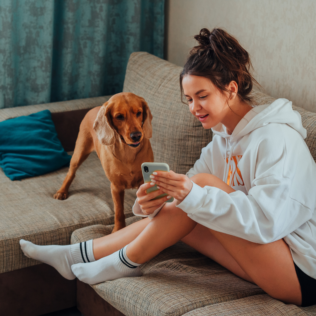 A woman is sitting on a couch looking at her phone next to a dog.