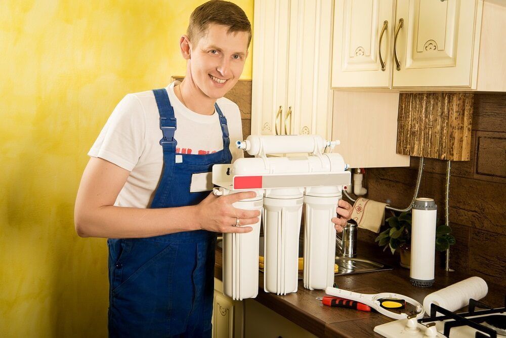 A Man is Standing in a Kitchen Holding a Cup of Water and a Water Filter — Brett Negus Plumbing In Hervey Bay, QLD