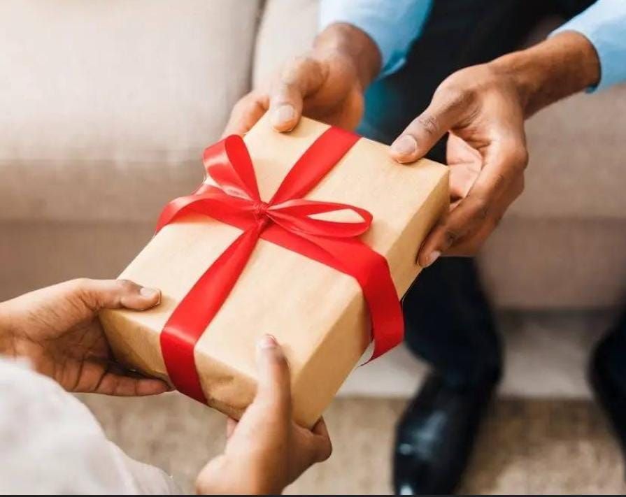 Manos intercambiando un regalo envuelto en papel marrón con una cinta roja; entorno interior.