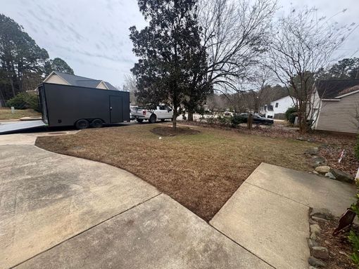 Curved concrete driveway beside a leaf-covered yard, with a tree, parked trailer, and houses on a cloudy day