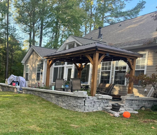 Covered patio with wooden beams and stone walls on a house, overlooking a grassy yard and trees