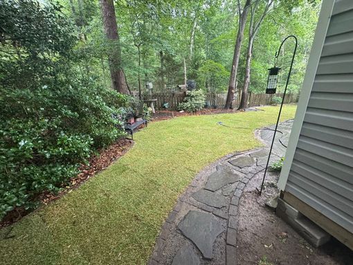 Backyard with green lawn, trees, stone walkway, and house siding on the right