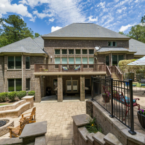 Backyard patio with brick house, balcony, stone seating area, and poolside fence under a blue sky