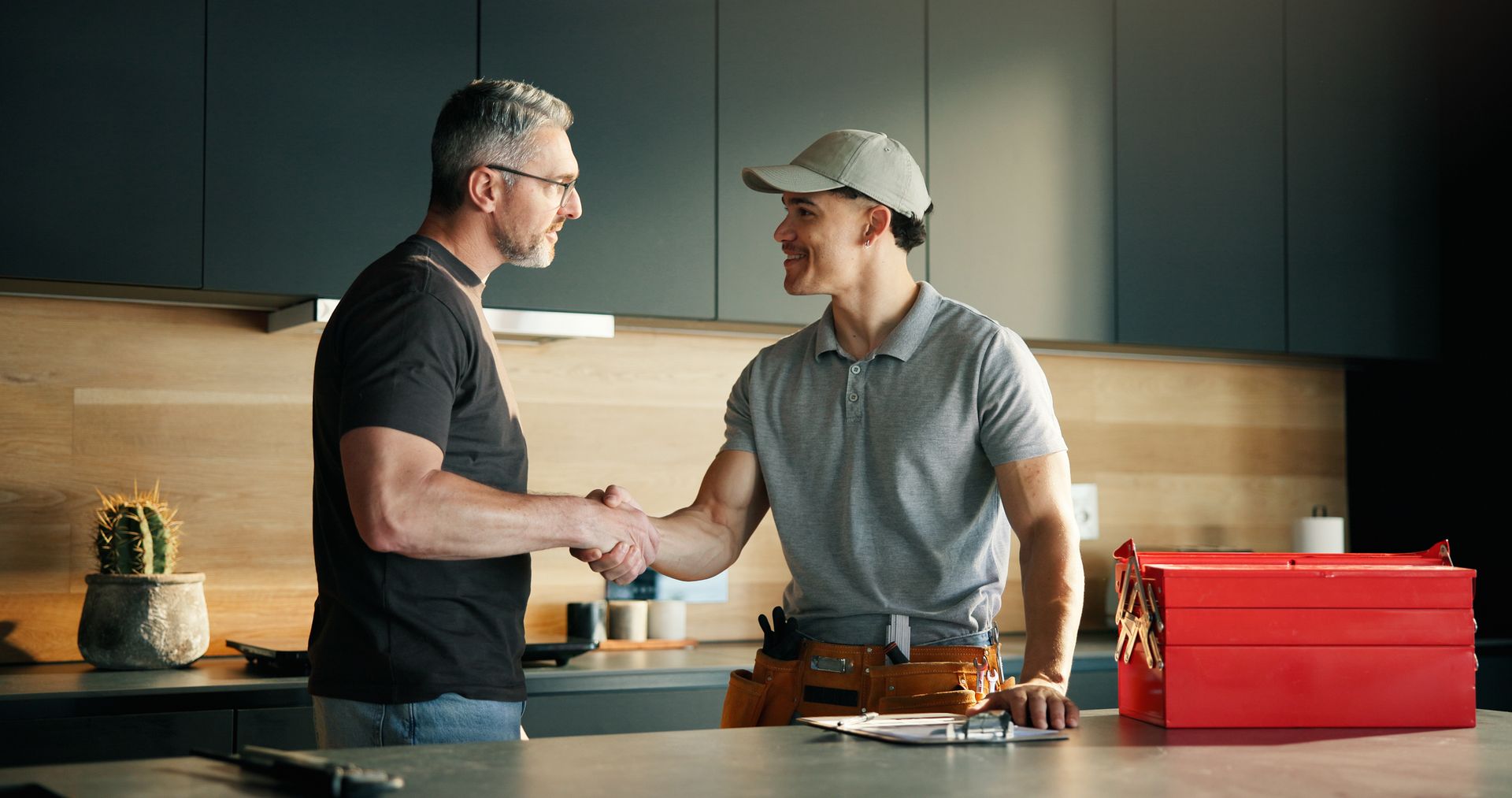 Two men shake hands in a kitchen beside a red toolbox and small tools on the counter.