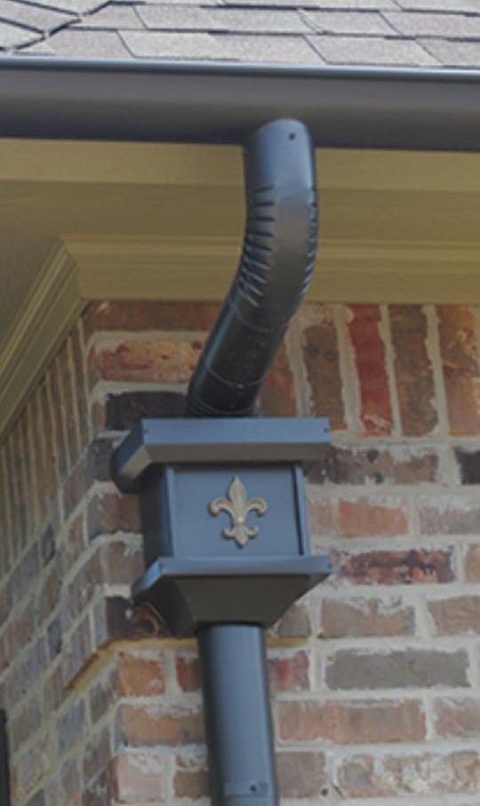 Dark gray downspout with fleur-de-lis detail, attached to a brick building with brown shingles.