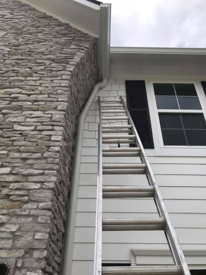 Ladder leaning against white house siding, near a stone chimney and window.