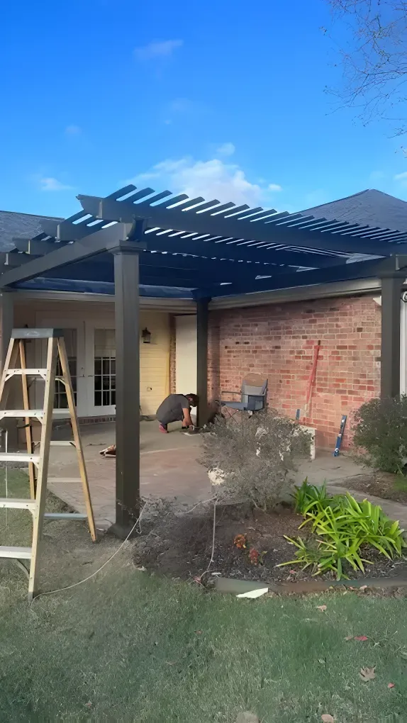 Pergola over patio, brown posts, dark blue beams, brick wall, ladder, plants, and blue sky.