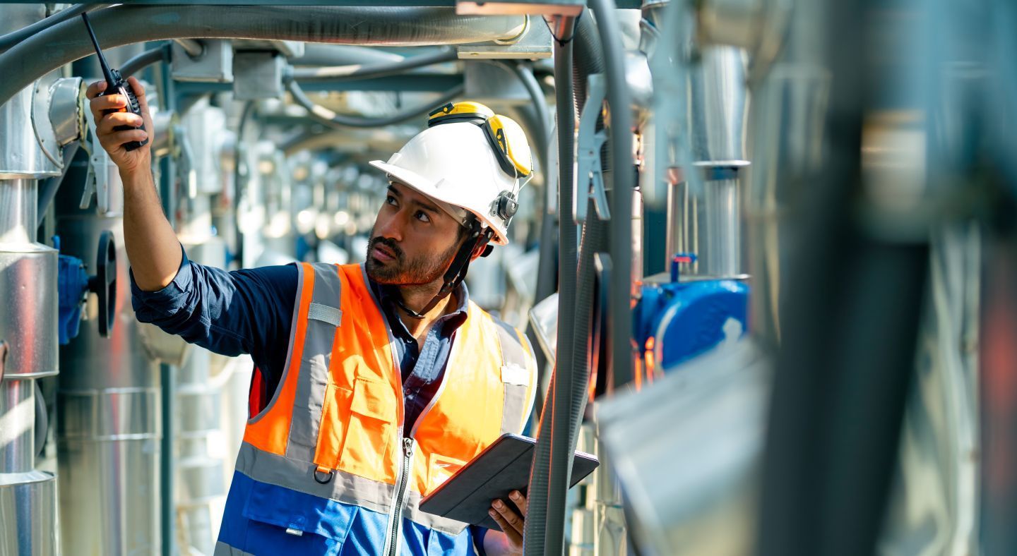 Worker in safety gear inspecting pipes with a tablet and radio in an industrial setting.