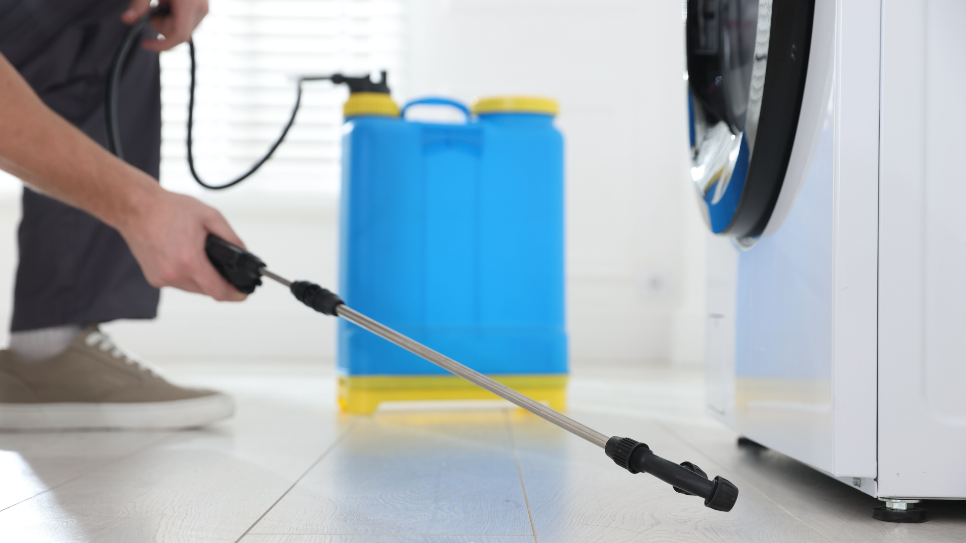 Person pressure-washing floor near a blue detergent tank beside a white washing machine.