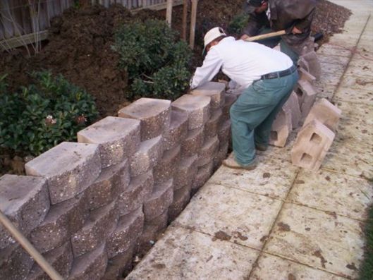 Worker in white shirt and green pants stacking gray retaining wall blocks along a sidewalk