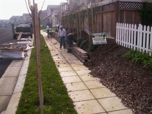 Workers repairing a sidewalk beside a road, with a fence, grass strip, and utility pole.