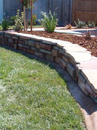 Curved stacked-stone retaining wall bordering a green lawn and landscaped garden bed