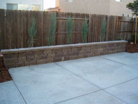 Concrete patio with a low stone retaining wall and wooden fence in a backyard