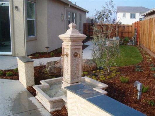 Decorative stone fountain in a landscaped front yard beside a house and wooden fence