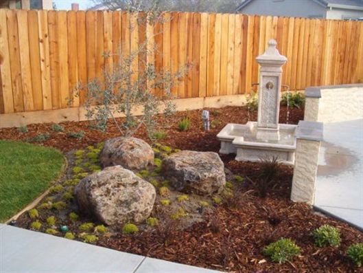 Landscaped backyard with large rocks, mulch, a small tree, and a decorative stone fountain beside a fence