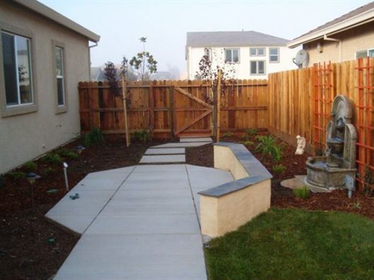 Backyard patio with concrete walkway, curved bench, wooden fence, and gate between two houses