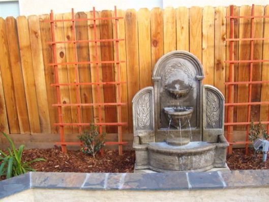 Stone garden fountain in front of a wooden fence with orange trellises and red mulch