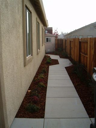 Narrow concrete walkway between a stucco house and wooden fence with flower beds along the side