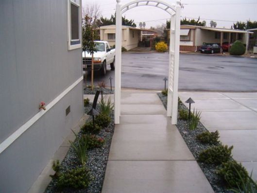 Narrow concrete walkway beside a building, leading through a white archway to a parking lot and homes beyond.