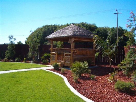 Thatched gazebo in a landscaped garden with green lawn and flower beds under a blue sky