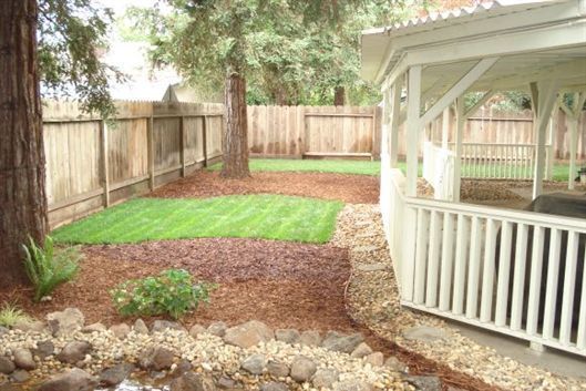Backyard with green lawn, stone borders, wooden fence, and white gazebo or porch on the right