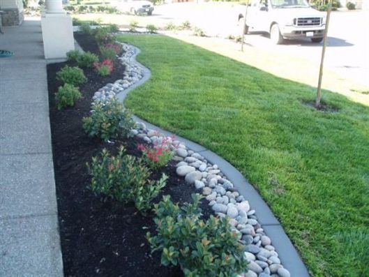 Curved landscaped garden bed with black mulch, white rocks, shrubs, and a manicured lawn beside a sidewalk