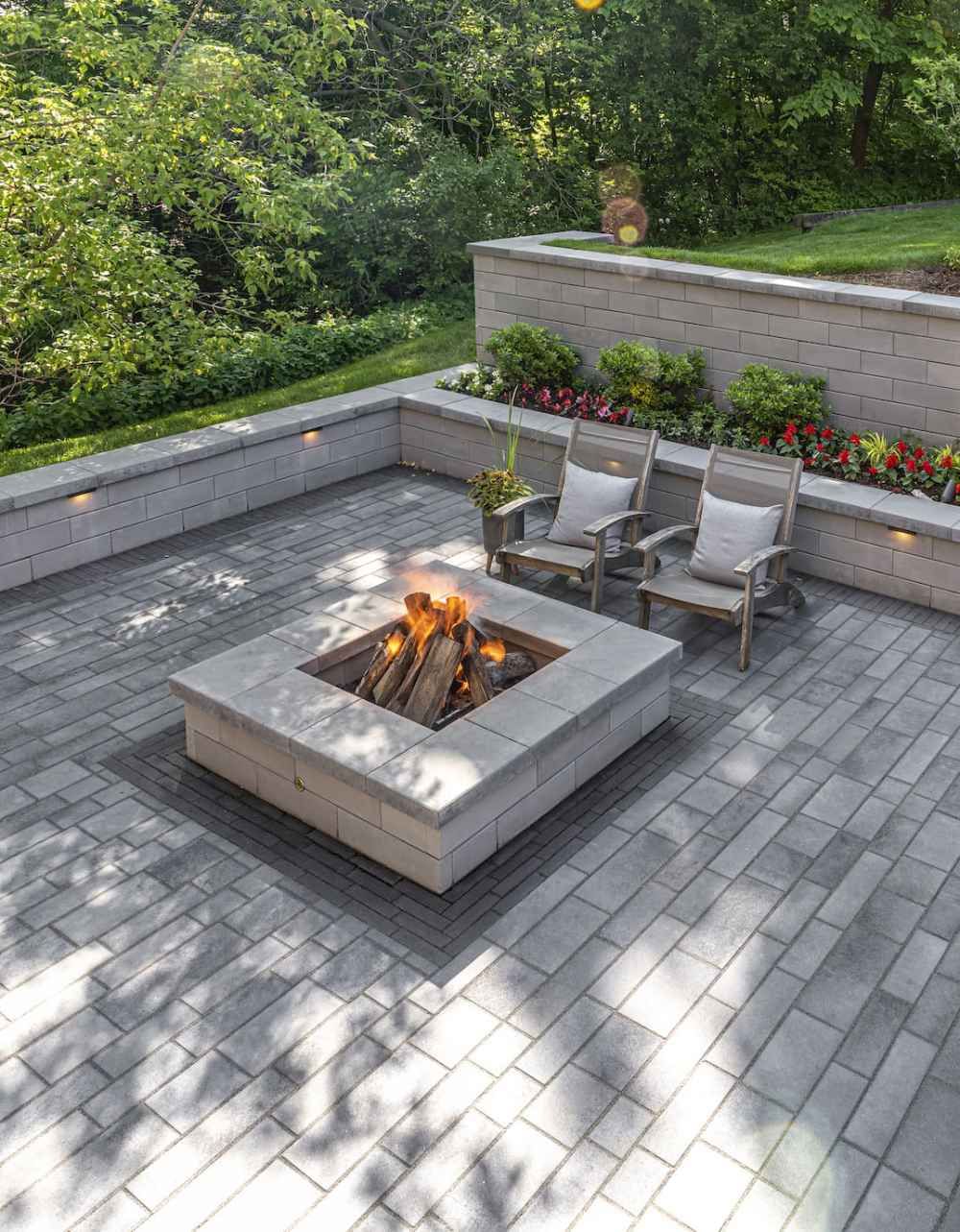 Modern stone patio with a square fire pit, two chairs, and flower planters surrounded by greenery