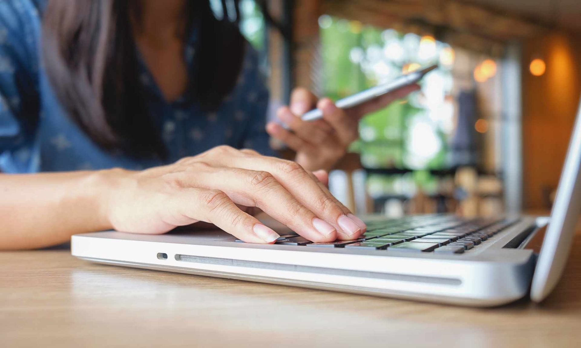 A person typing on a laptop computer while holding a smartphone in their other hand at a table.