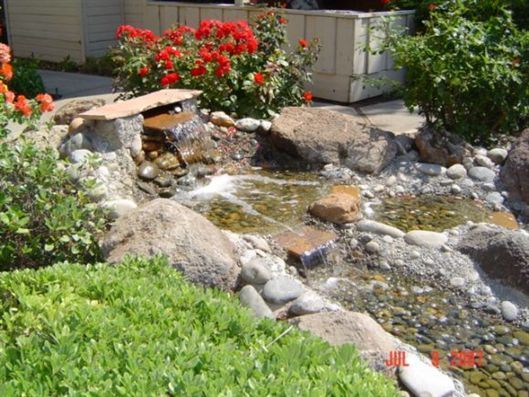 Small backyard pond with flowing water, rocks, and bright red flowers beside green plants