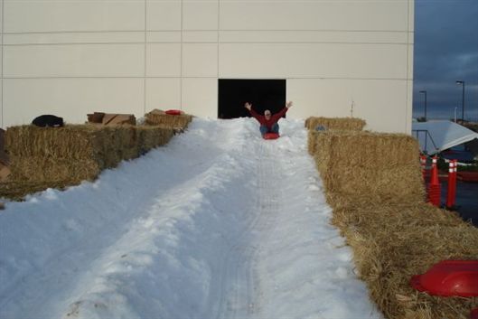 Person sledding down a snowy ramp bordered by hay bales toward a building entrance
