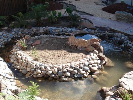 Rock-lined garden pond with a small stone waterfall and sandy island, surrounded by water and plants