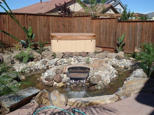 Backyard pond with rocks and waterfall, wooden fence, and a light wood hot tub behind it