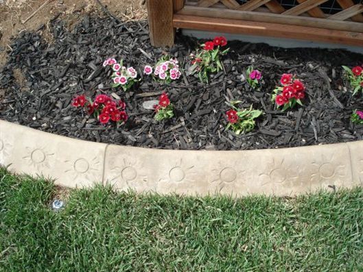 Flower bed with red and pink flowers along a curved stone border beside grass and mulch.