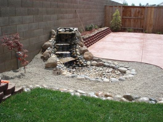 Backyard with a small rock waterfall, gravel bed, green lawn, and concrete patio by a wooden fence