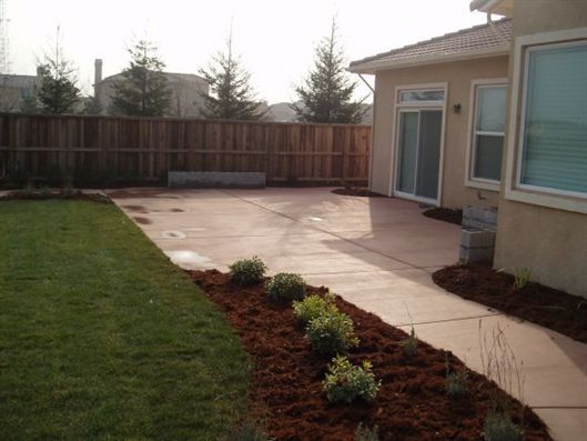 Backyard patio with concrete slab, lawn, flower bed, wooden fence, and house exterior