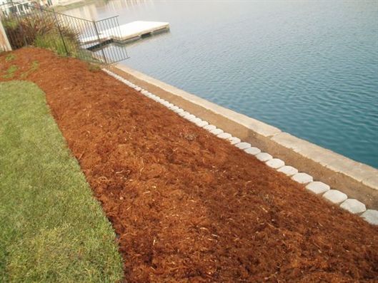 Landscaped lakeside slope with red mulch, grass, and a white rock border beside calm blue water