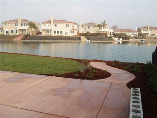 Waterfront residential homes across a calm canal, with a curved walkway and lawn in the foreground.