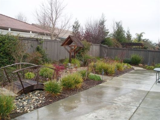 Rainy backyard garden with wet patio, small bridge, bird feeder, and fenced landscaping beds