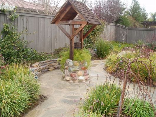 Backyard garden path with a small wooden wishing well, stone border, and shrubs along a fence