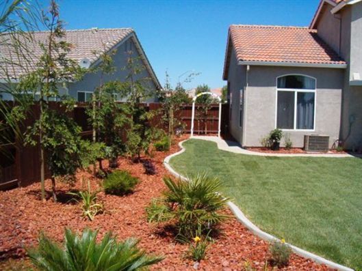 Suburban backyard with green lawn, red mulch, shrubs, and two houses under a clear blue sky
