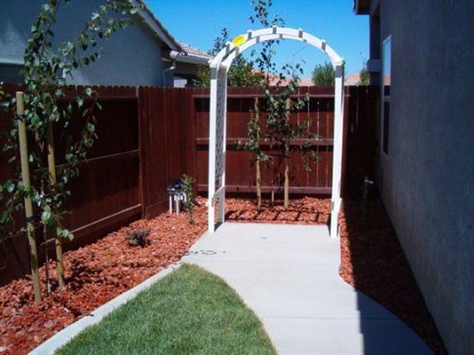 Narrow backyard path leading to a white garden arch beside a red fence and mulched beds