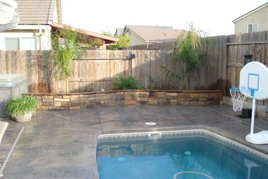 Backyard patio with a small swimming pool, stone wall, wooden fence, and a basketball hoop